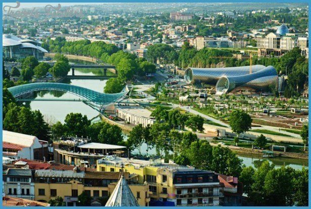 Panoramic view of Tbilisi, Georgia, with the Peace Bridge, buildings, and river Kura. Lush greenery and sunny weather.