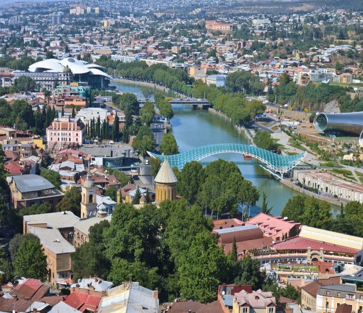 Aerial view of Tbilisi, Georgia, with buildings, a river, and a bridge. Sunny day with a mix of old and modern architecture.