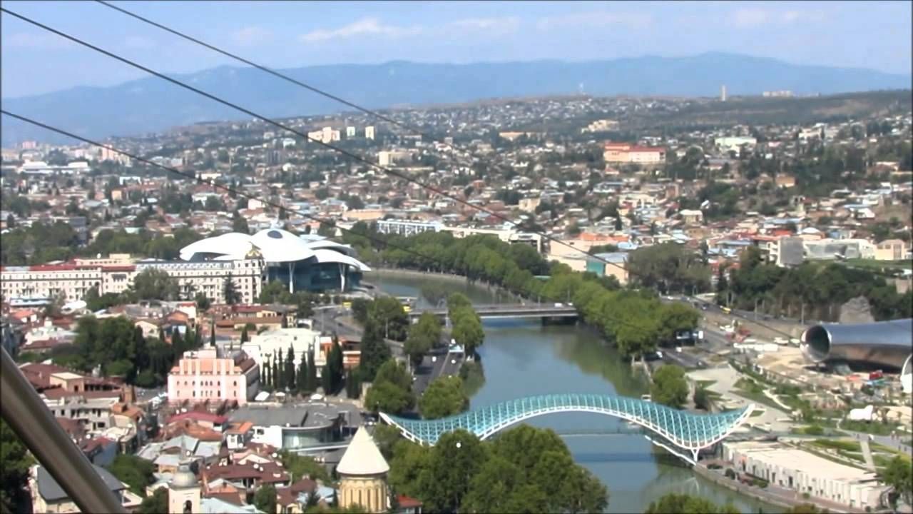 A cityscape of Tbilisi, Georgia, with a river, bridges, and a cable car line. Buildings of various colors are spread across a valley.