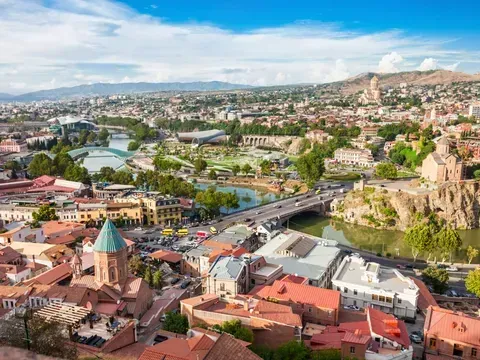 Aerial view of Tbilisi, Georgia, with red-roofed buildings, a river, bridges, and a distant mountain under a blue sky.