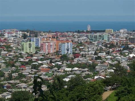 Cityscape with colorful buildings, trees in the foreground, and the sea in the distance under a blue sky.