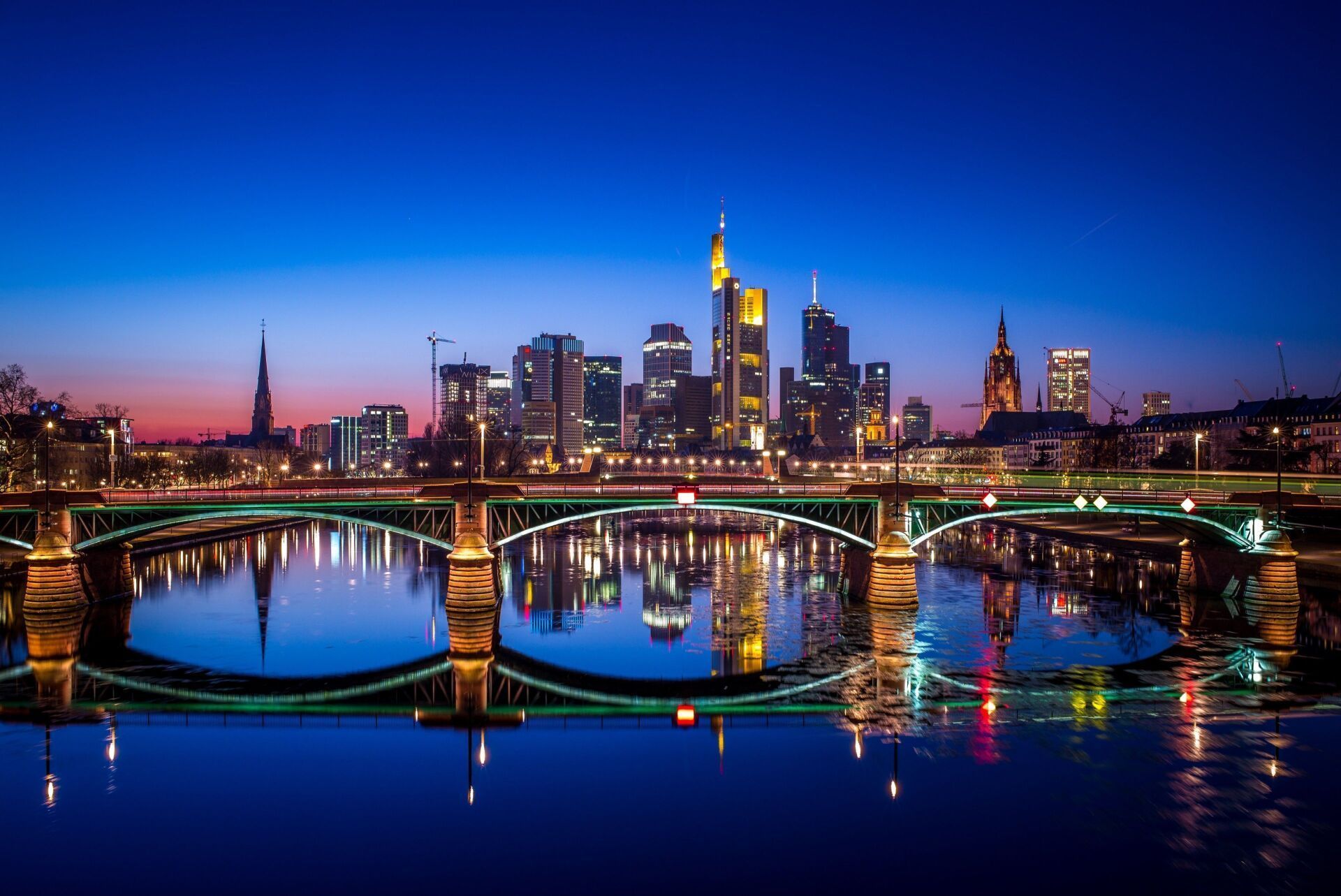 Frankfurt skyline at dusk, reflected in the Main River; city lights illuminating the bridge and buildings against a dark blue sky.