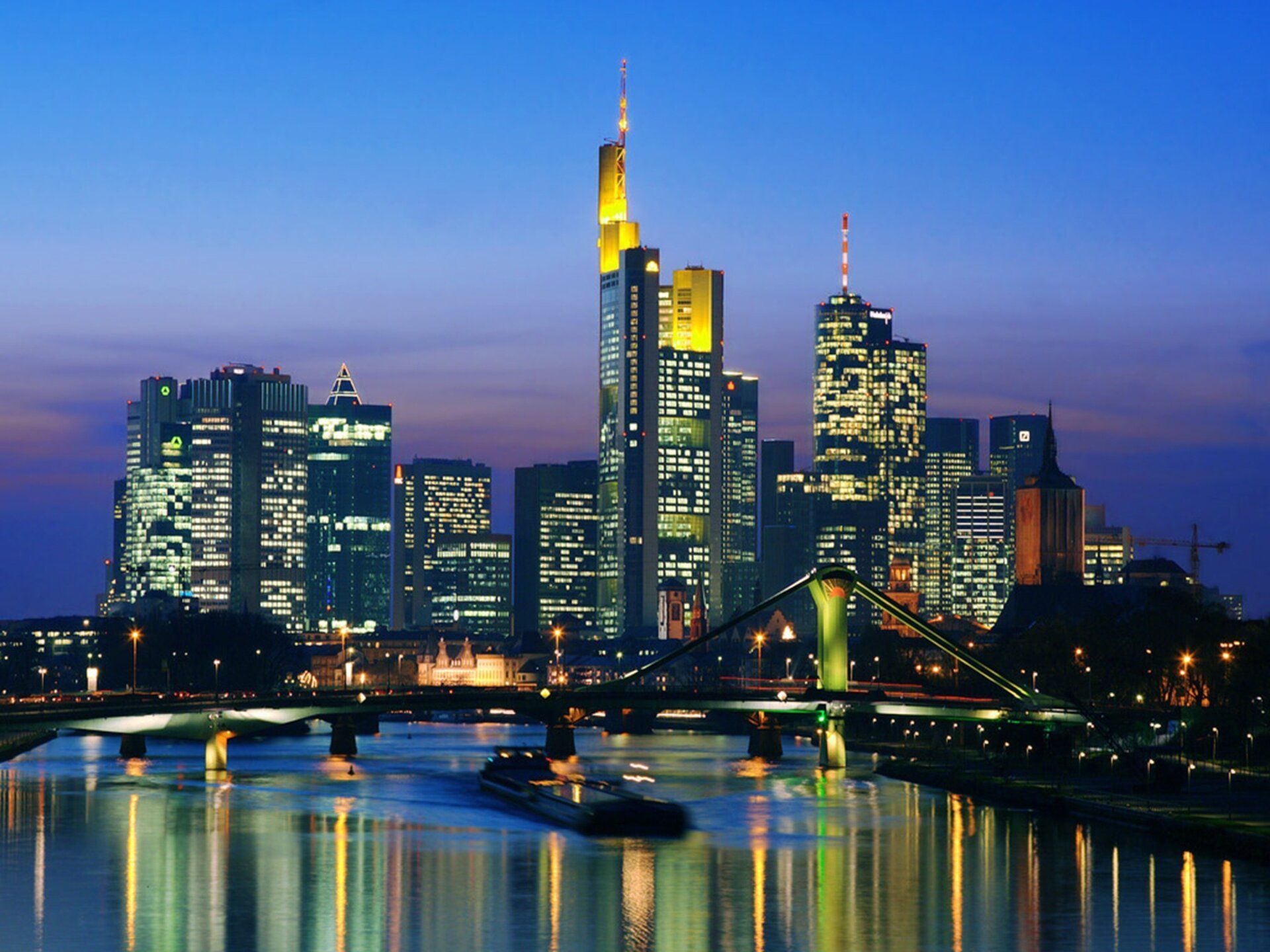 Frankfurt skyline at dusk, with illuminated skyscrapers reflecting in the Main River. A bridge is visible in the foreground.