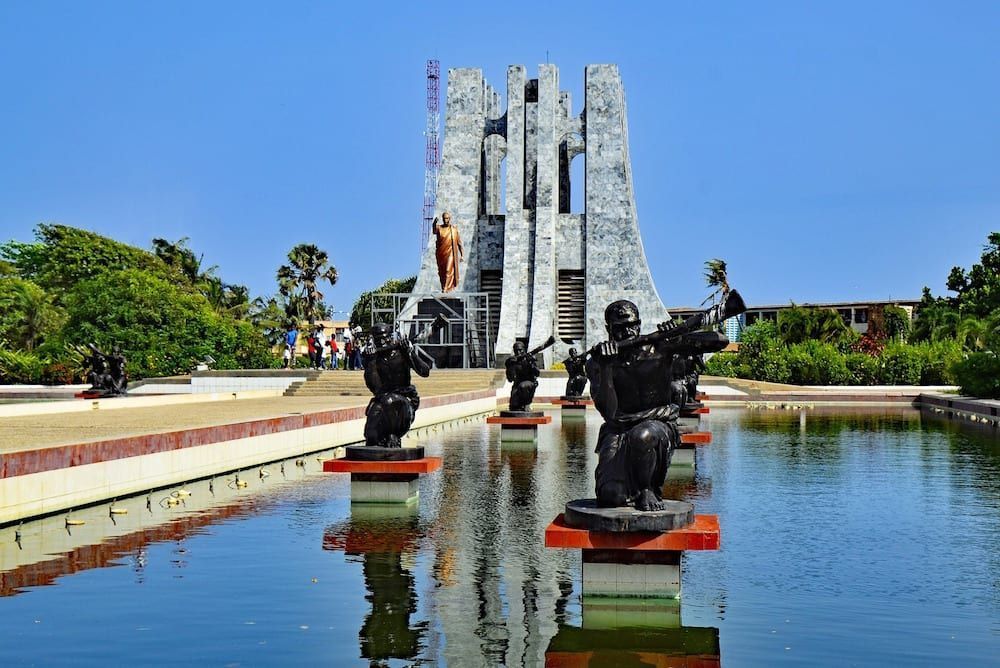 Kwame Nkrumah Mausoleum in Accra, Ghana, with bronze statues in a reflecting pool. The monument has a tall, open-air structure with a statue visible.