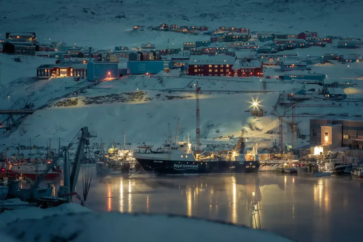 A snowy harbor at night with a ship docked, surrounded by buildings lit up on a hillside. The scene has a cold, blue-toned aesthetic.