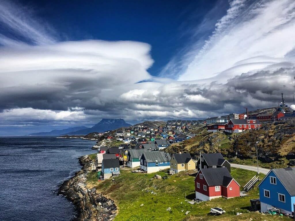Colorful houses line a rocky coast in Greenland, under a dramatic sky with swirling clouds.