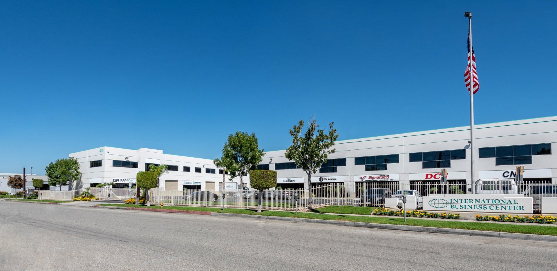 A large, white commercial building under a clear blue sky. An American flag flies on a pole. A sign is visible.