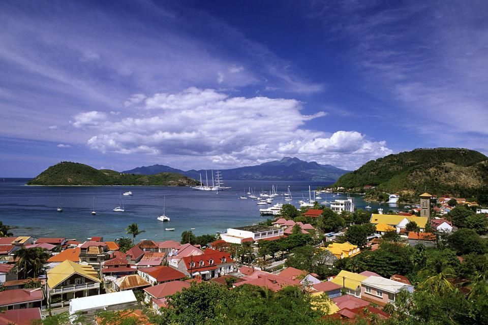 Coastal village with colorful houses, sailboats in a harbor, green hills, and a blue sky with clouds.
