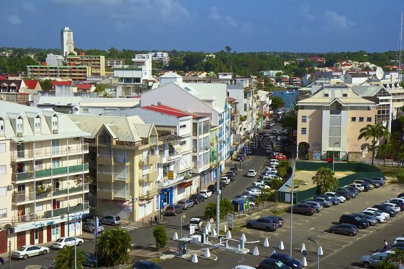 A cityscape with buildings of various heights and colors, a street lined with parked cars, and a tall white tower in the distance.