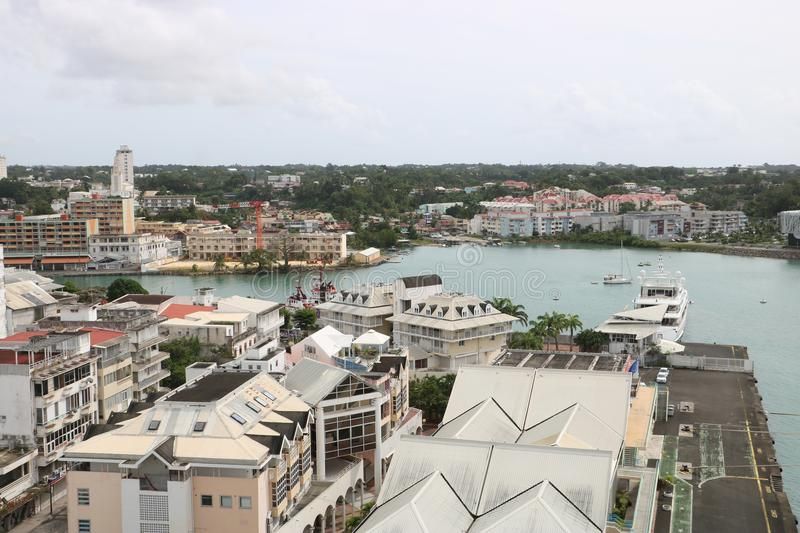 View of a waterfront city with buildings, harbor, boats, and lush green trees under a cloudy sky.