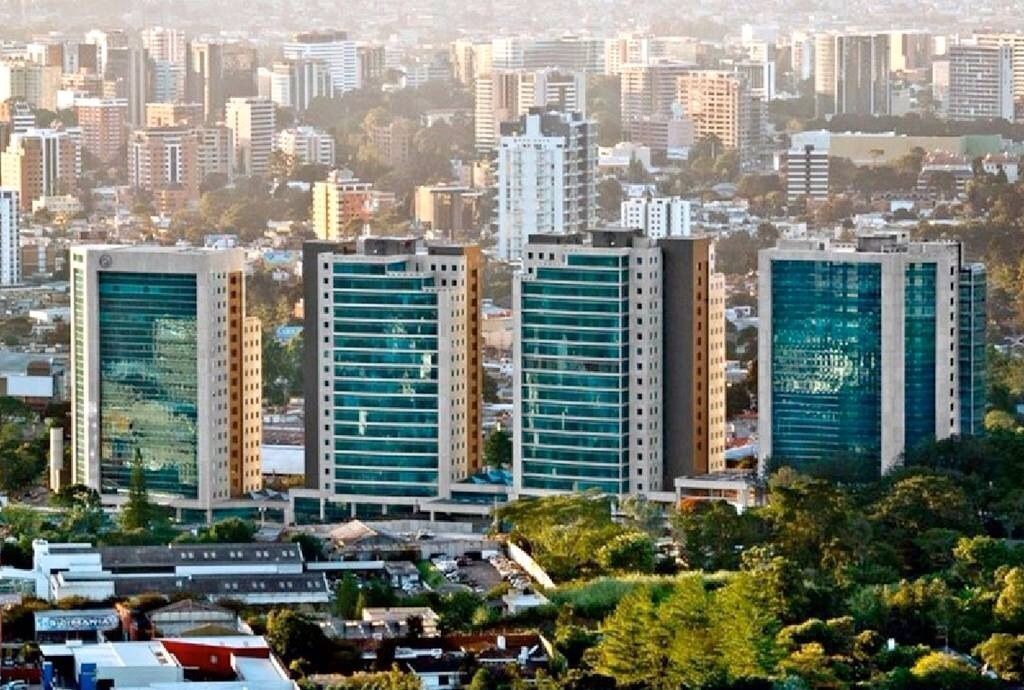 Four modern high-rise buildings with teal glass facades in an urban setting, surrounded by buildings and trees.