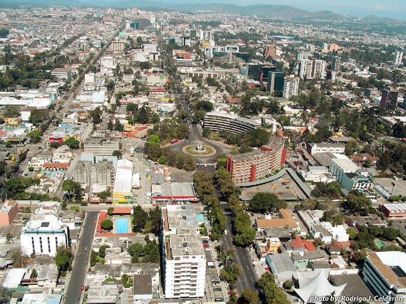 Aerial view of a city with long streets, buildings, and a roundabout with trees at the center.