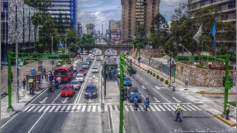 Busy city street scene in Guatemala City. Cars and pedestrians cross at a marked crosswalk with high-rise buildings in the background.