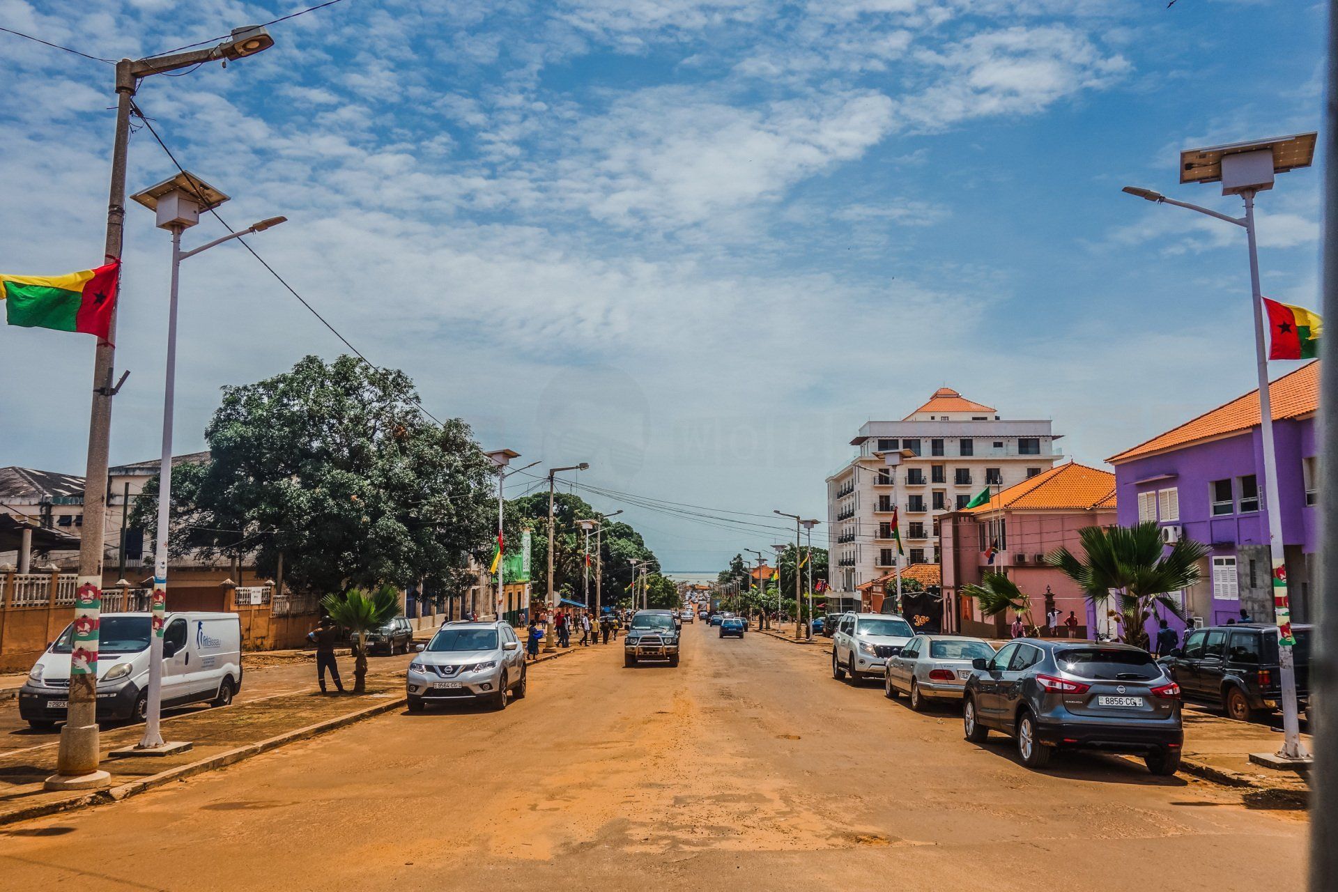 Street scene in Guinea-Bissau. Cars driving on a dirt road flanked by buildings and streetlights with solar panels. National flags fly overhead.