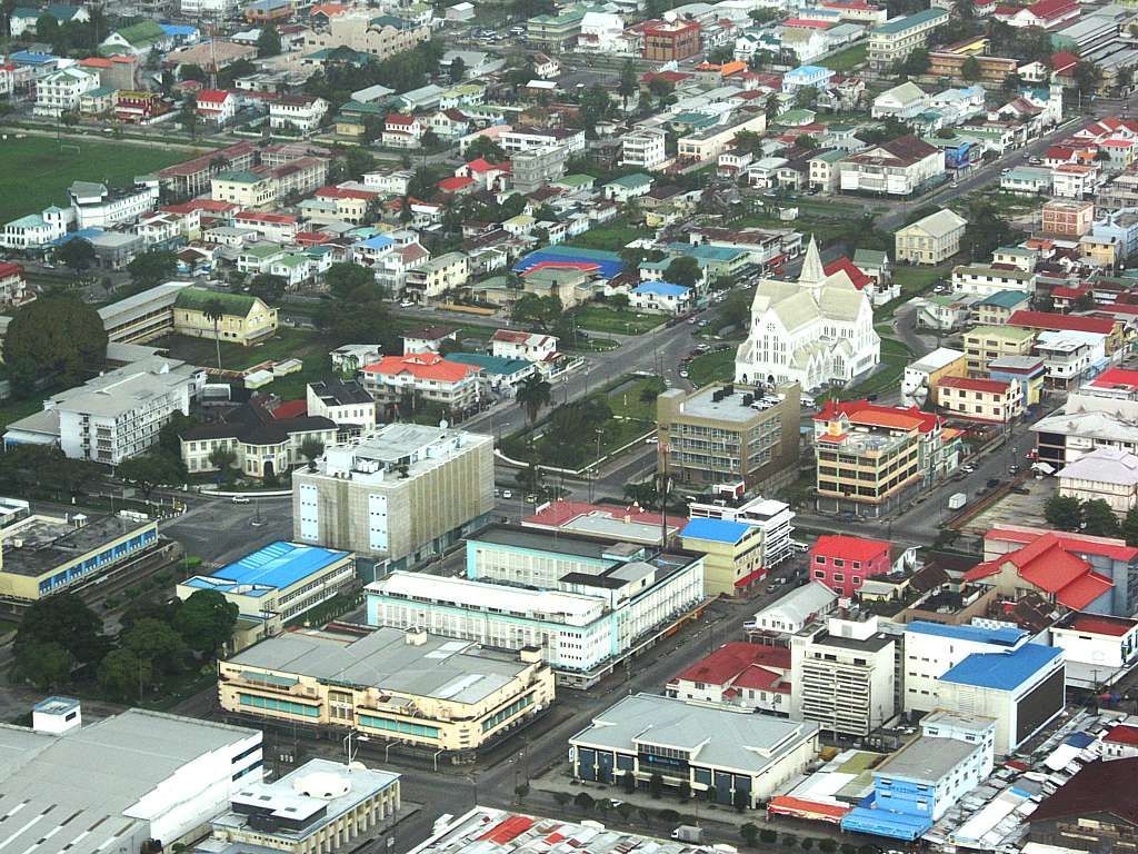 Aerial view of a city with colorful buildings and roads. Notable structures include a large white building and several buildings with blue roofs.