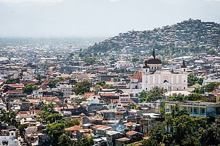 A sprawling cityscape with a large white church in the center, a hillside densely populated with houses, and a hazy sky.