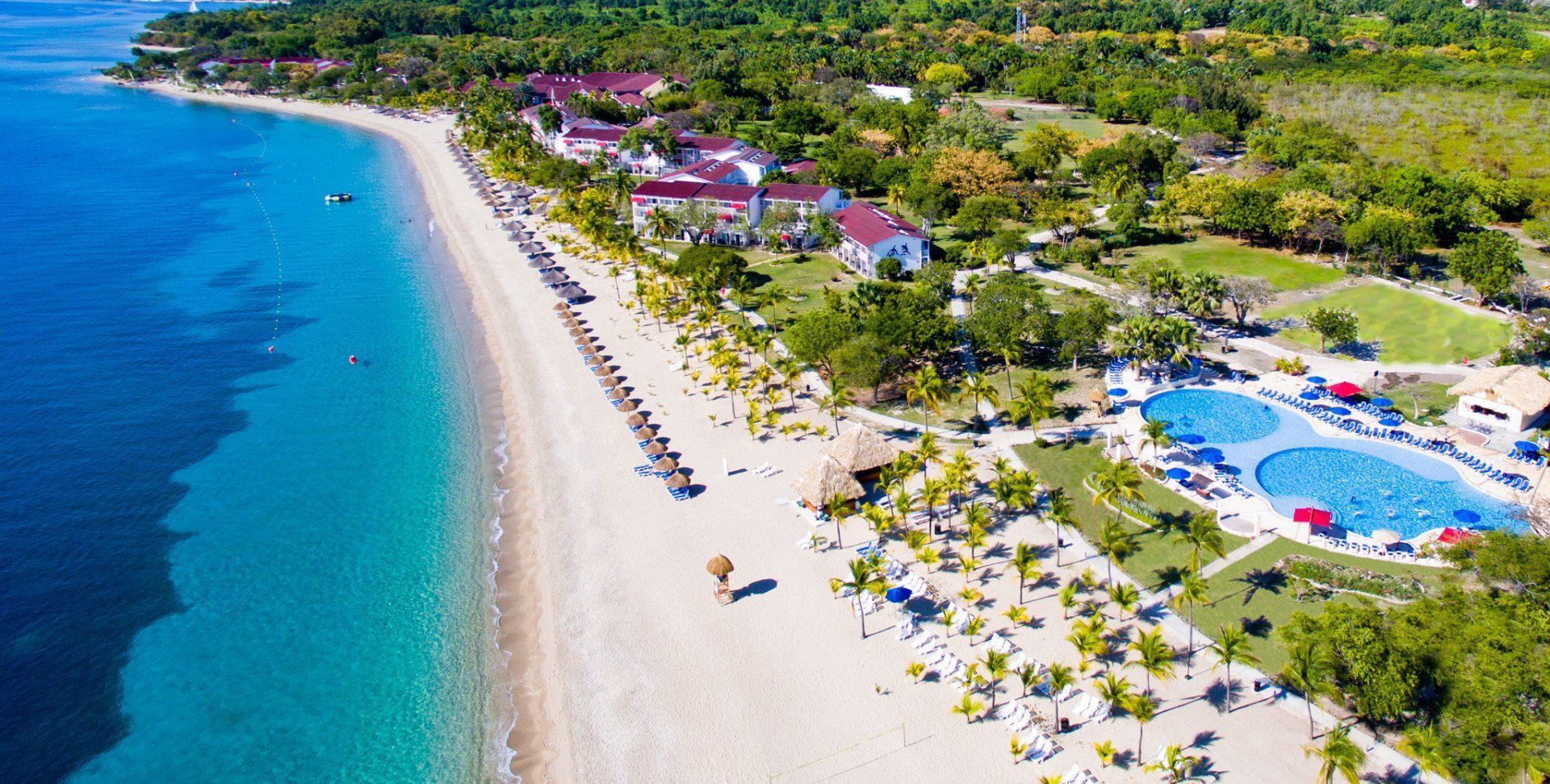 Aerial view of a beachfront resort with a pool, sandy beach, turquoise water, and resort buildings.