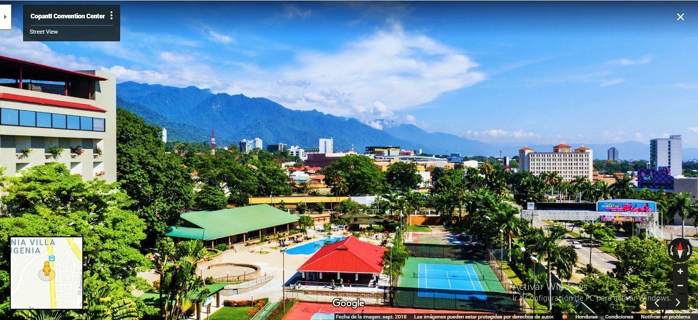 A cityscape with lush greenery, buildings, tennis courts, and mountains under a bright blue sky.