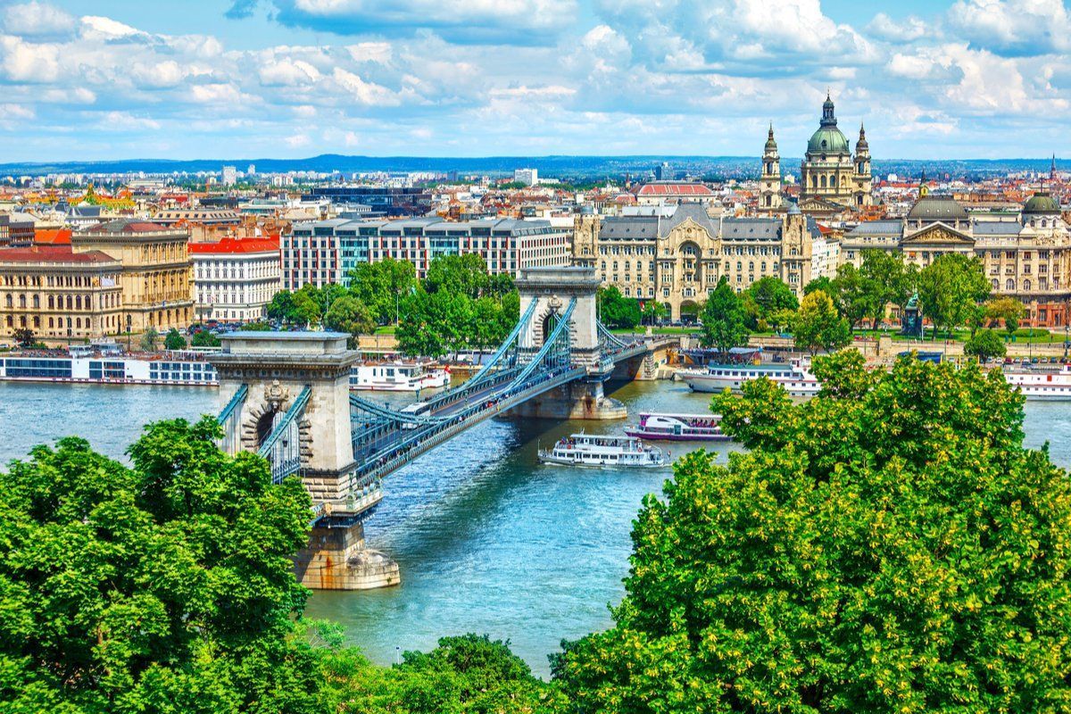 A picturesque view of Budapest with the Chain Bridge over the Danube River, lush green trees, and buildings under a blue sky.
