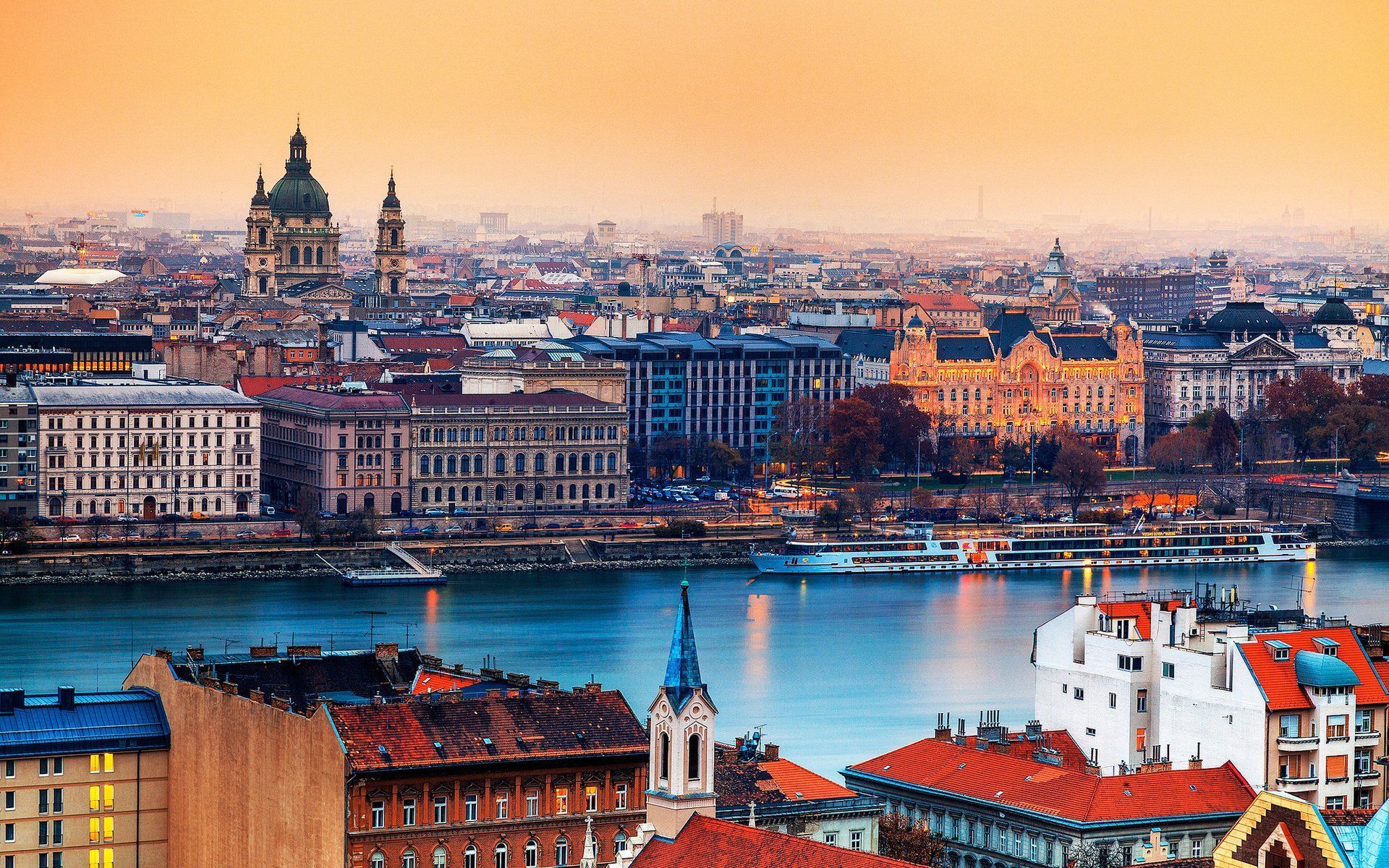 Budapest skyline at sunset with buildings lining the Danube River; includes St. Stephen's Basilica and other historical architecture.