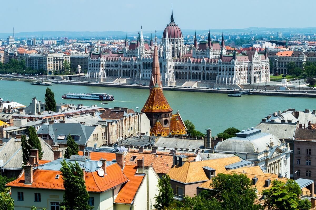 A cityscape of Budapest, Hungary, with the Danube River, Parliament Building, and various buildings with orange rooftops under a clear blue sky.