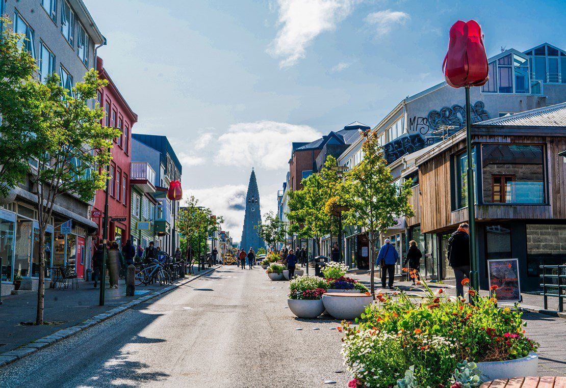 Street scene in Reykjavik, Iceland, with colorful buildings, people walking, and a prominent church tower in the distance.