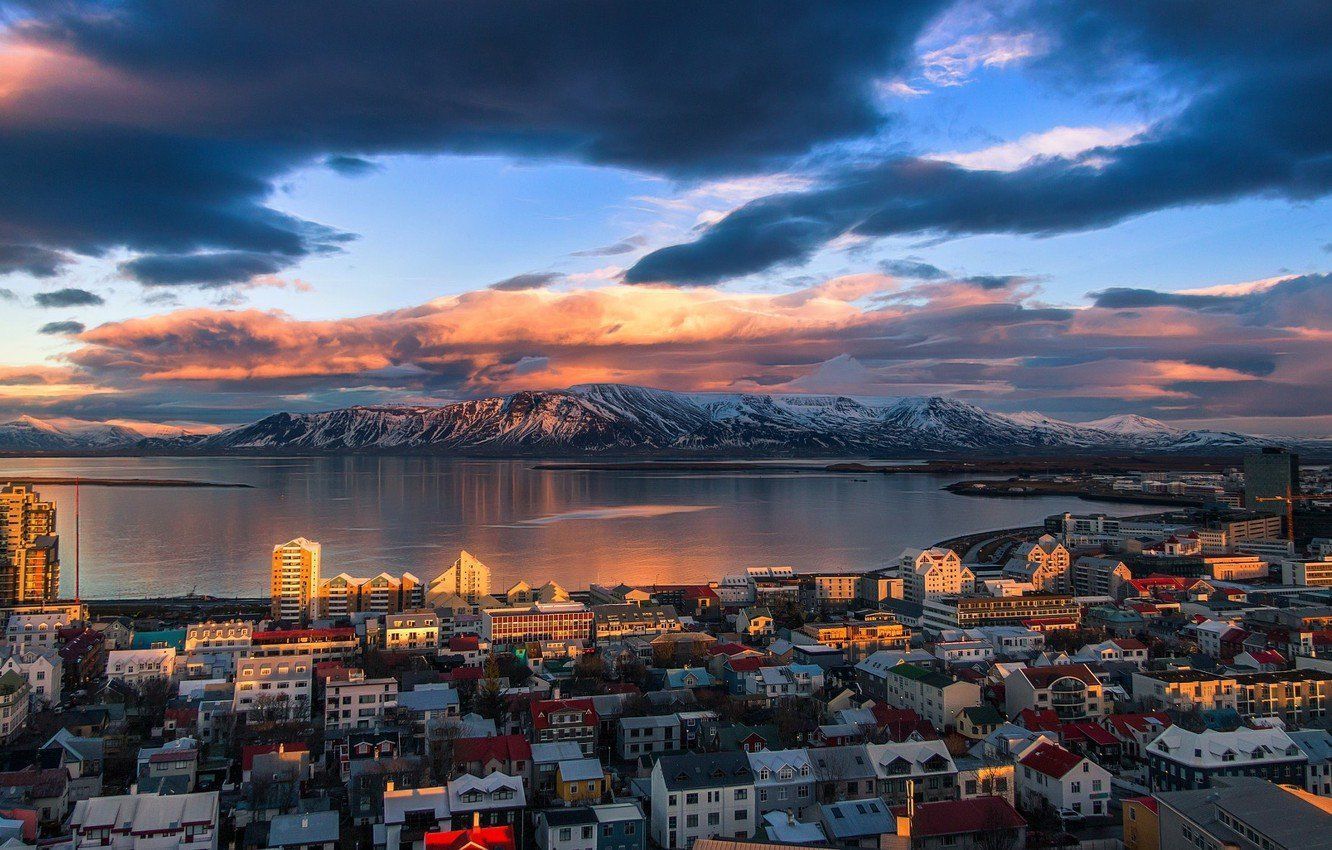 Cityscape at sunset with a body of water and snow-capped mountains in the background, under a vibrant cloudy sky.