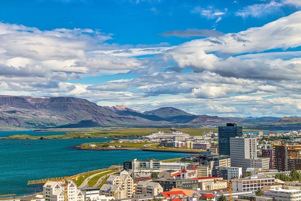 A cityscape with buildings and a blue body of water against a backdrop of mountains under a cloudy sky.