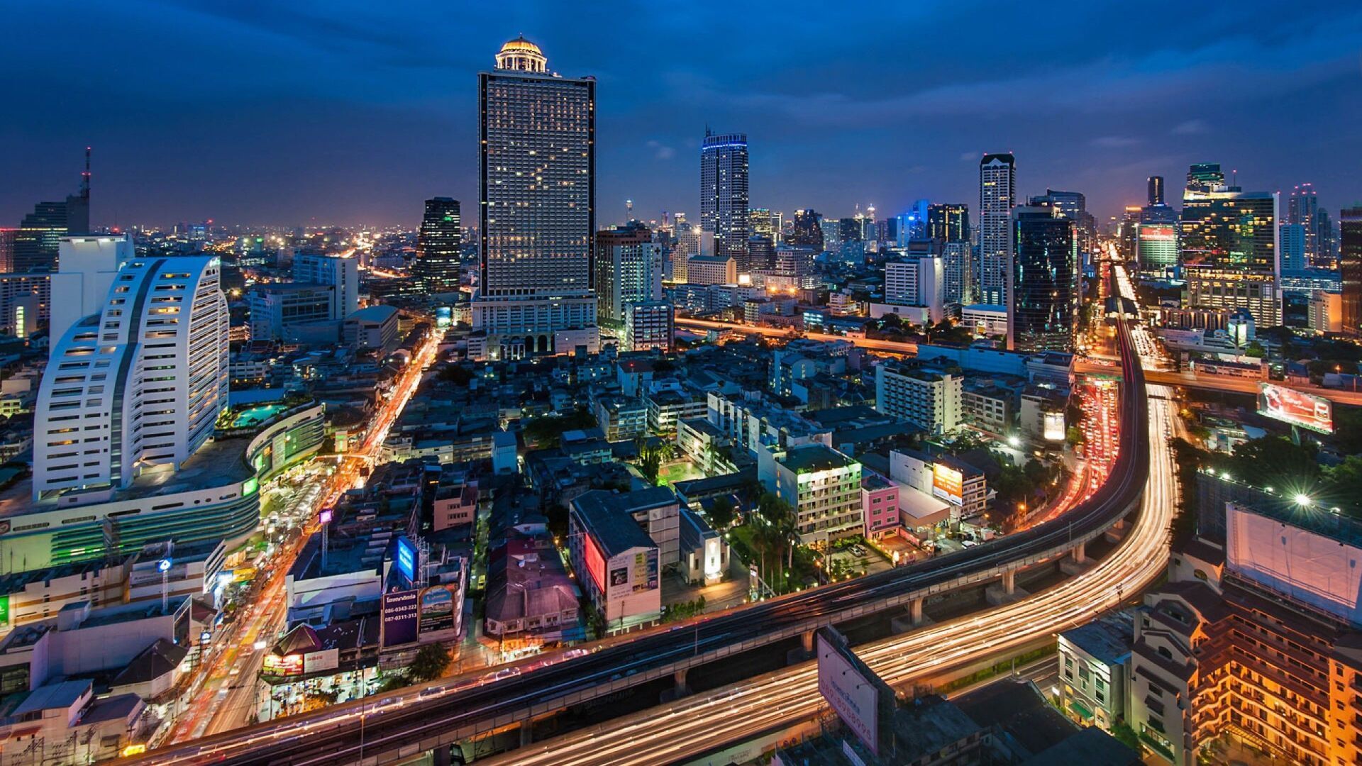 Night view of a brightly lit cityscape, with skyscrapers and busy roadways. Blue sky backdrop with city lights reflecting.