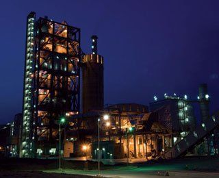 Industrial plant illuminated at night, featuring a tall, metal-framed structure and several other buildings against a dark blue sky.