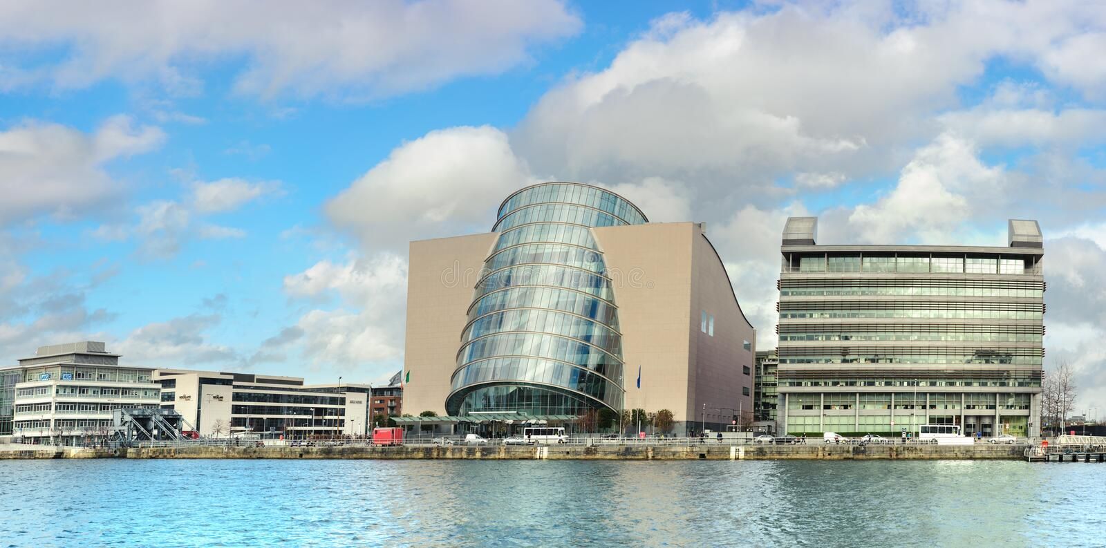 Modern buildings line a waterfront under a cloudy blue sky. The glass facade of the Convention Centre Dublin is prominent.