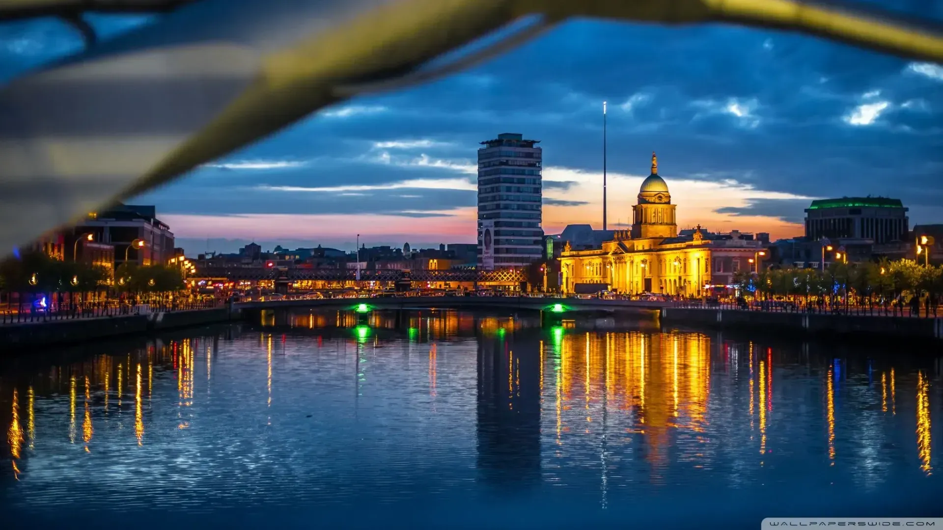 City skyline at dusk, reflected in a river. Lit buildings and bridge create a colorful scene with water reflections.