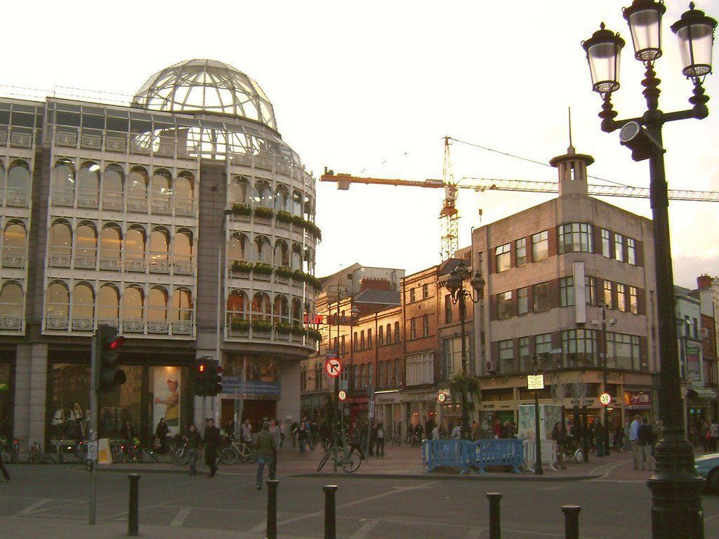 Busy Dublin street scene with shops, a crane in the background, and pedestrians at dusk.