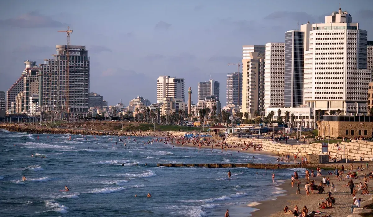 Beach scene in Tel Aviv, Israel, with people sunbathing and swimming. Tall buildings line the shore under a blue sky.