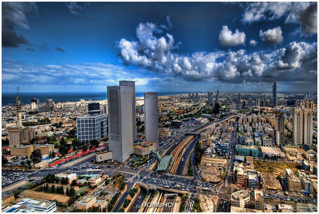 Tel Aviv skyline with tall buildings, city streets, and the Mediterranean Sea under a blue sky with clouds.