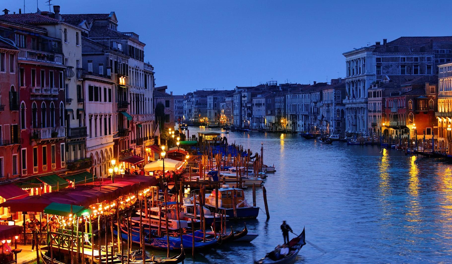 A nighttime view of the Grand Canal in Venice, Italy, with brightly lit buildings and gondolas on the water.