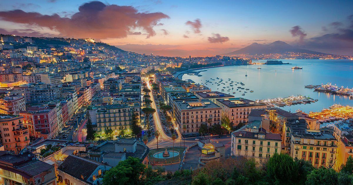 Cityscape of Naples, Italy at dusk. Buildings line the coast with lights illuminating the water, bay, and mountain in the background.