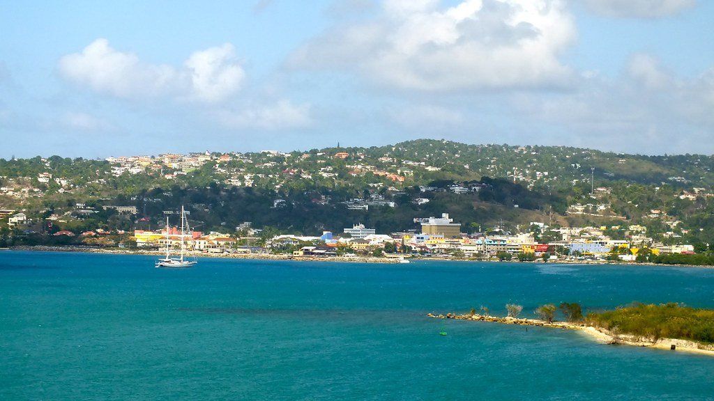 Coastal cityscape with turquoise water, a sailboat, and a hilly, green landscape dotted with buildings under a partly cloudy sky.