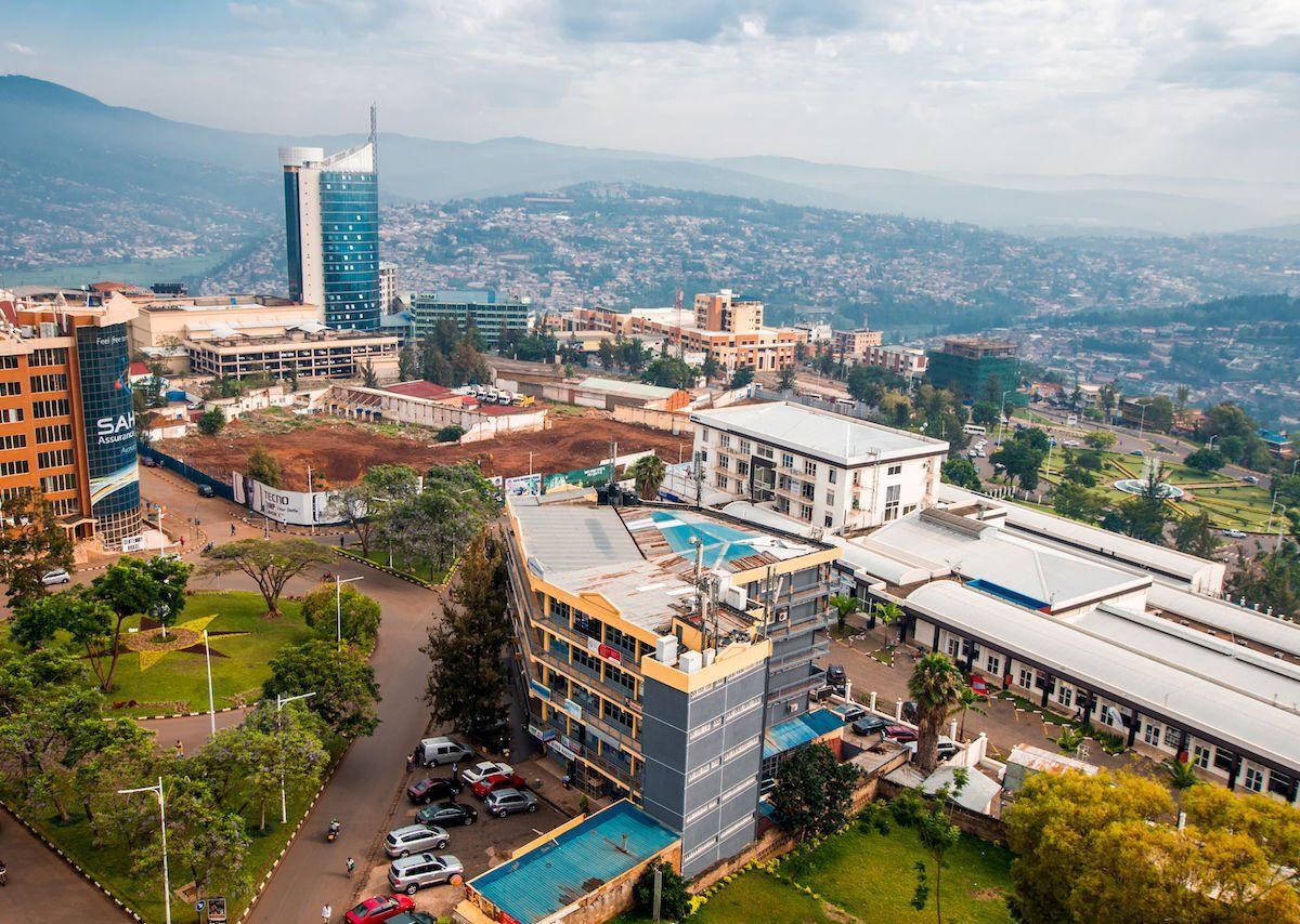 Aerial view of Kigali, Rwanda, with a tall skyscraper in the distance. Buildings and greenery fill the foreground with a hazy mountain range.