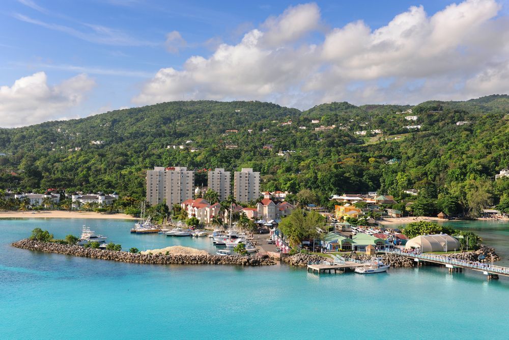 An aerial view of a coastal town with turquoise water, green hills, buildings, and boats docked at a harbor.