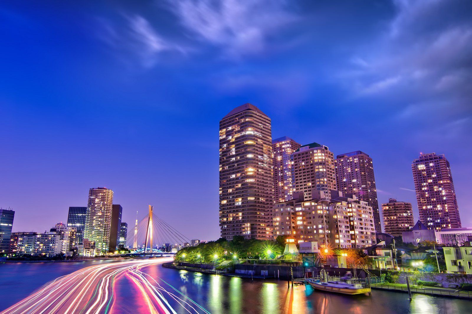 City skyline at dusk with illuminated skyscrapers, a river with light trails, and a bridge.