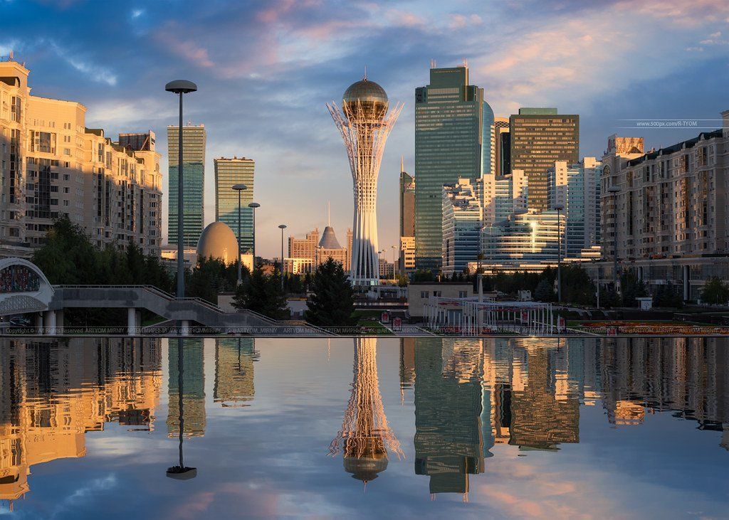A cityscape in Astana, Kazakhstan, reflecting in calm water. Buildings of varying heights and colors are set against a sky with golden hues.