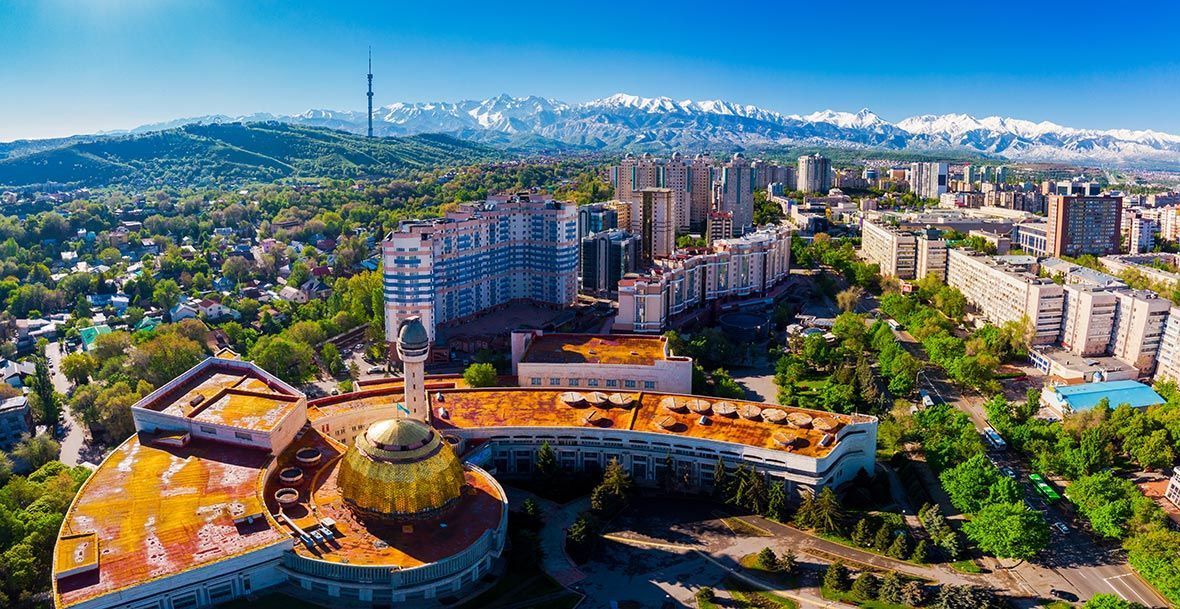 Aerial view of Almaty, Kazakhstan, showing urban landscape with residential buildings, trees, and mountains in the background under a clear blue sky.
