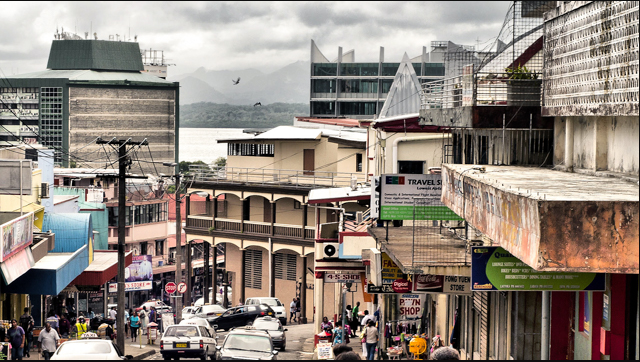 Busy street scene in Port Vila, Vanuatu, with shops, cars, people, and waterfront buildings under a cloudy sky.