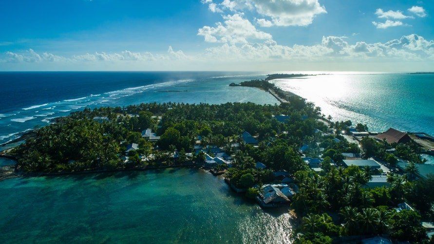 Aerial view of a tropical island surrounded by turquoise water and connected to a sandy spit; lush green vegetation and blue sky.