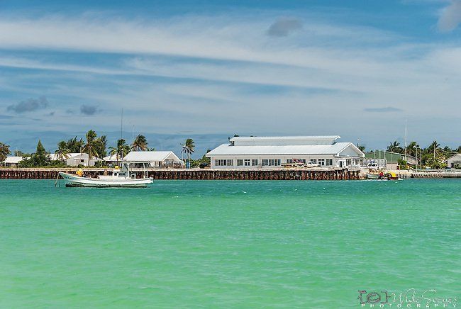 A white boat sails in turquoise water toward a pier with white buildings under a blue sky in a tropical location.
