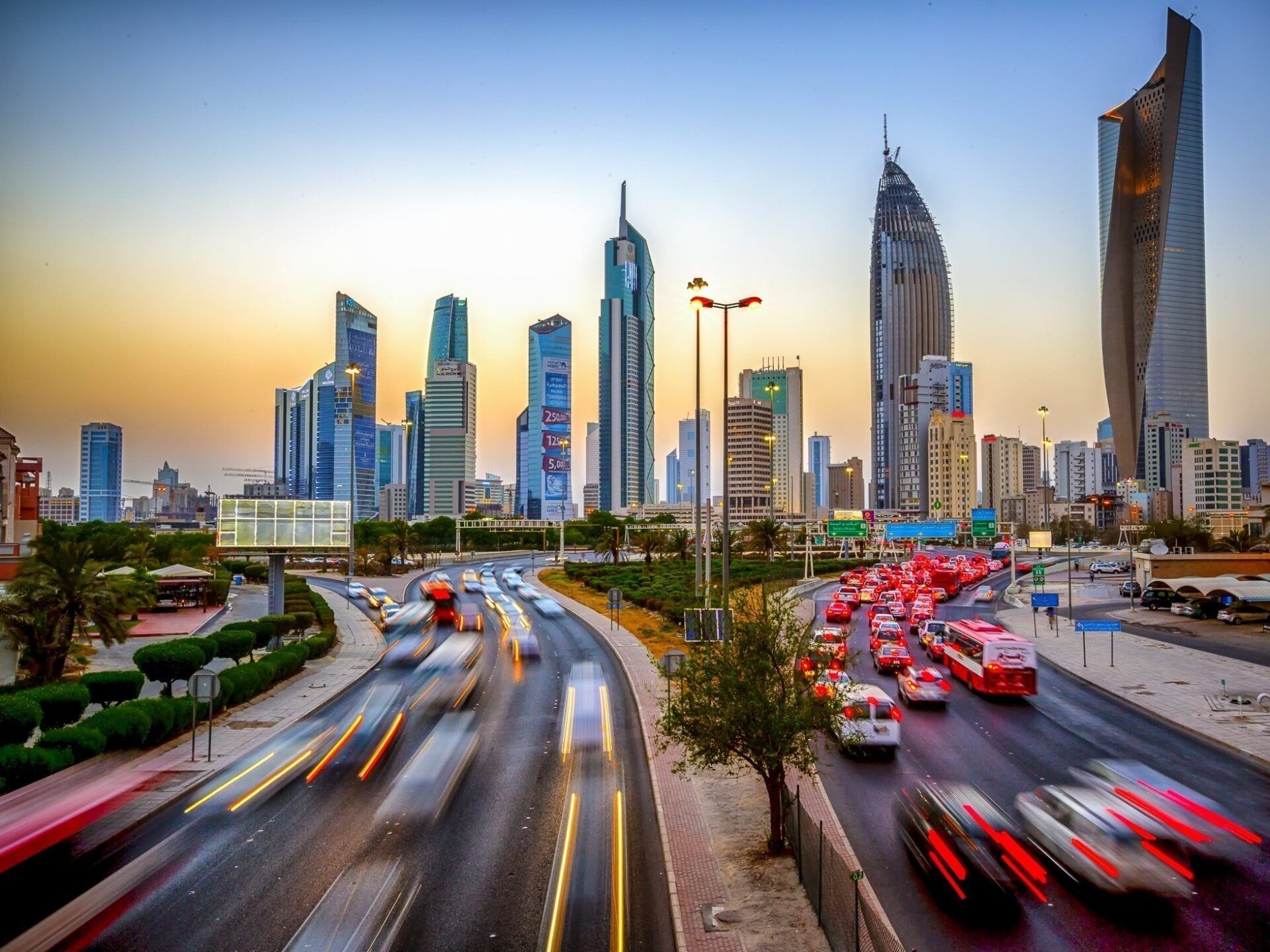 City skyline with skyscrapers silhouetted against a sunset, with a multi-lane road filled with blurred traffic.