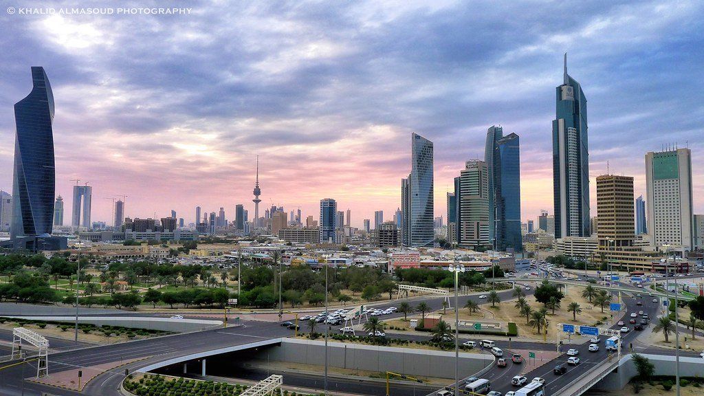 Kuwait City skyline at sunset with modern skyscrapers, roads, and the Kuwait Towers. Cloudy sky with pink and purple hues.
