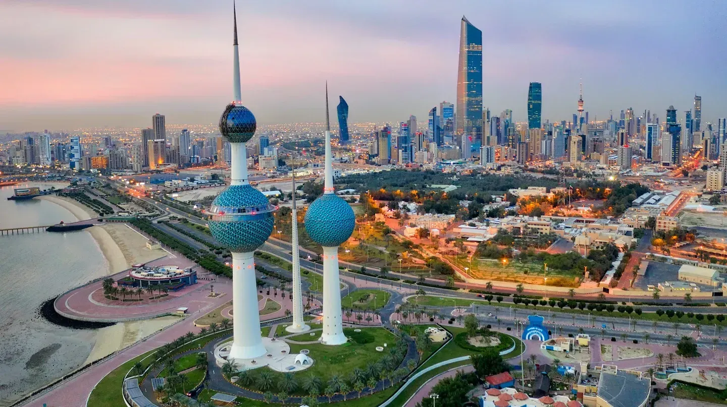 Aerial view of Kuwait City at dusk, featuring the iconic Kuwait Towers in the foreground and a modern skyline in the distance.