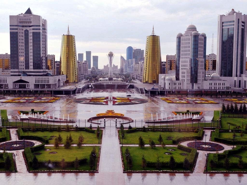 Cityscape of Astana, Kazakhstan, featuring modern skyscrapers, golden towers, and a central monument, reflecting in a large plaza with fountains and greenery.